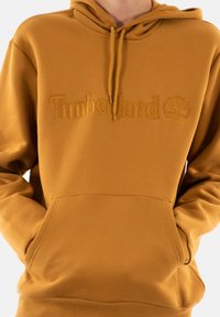 Felpa gialla senape con cappuccio, tasca a marsupio e logo "Timberland" in rilievo sulla parte frontale. Tessuto morbido e liscio.
