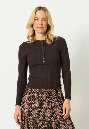 Blondynka ubrana w ciemny prążkowany top z długim rękawem i guzikami oraz czarną spódnicę w kwiatowy wzór, stojąca na jednolitym jasnym tle.