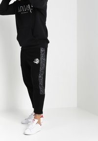 Černé tepláky s lesklým stříbrným pruhem a grafikou lebky, vyrobené z měkké tkaniny. Spárované s černou mikinou a bílými teniskami.