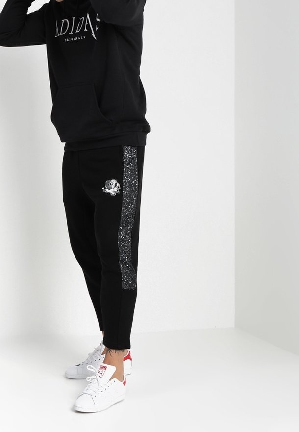 Černé tepláky s lesklým stříbrným pruhem a grafikou lebky, vyrobené z měkké tkaniny. Spárované s černou mikinou a bílými teniskami.