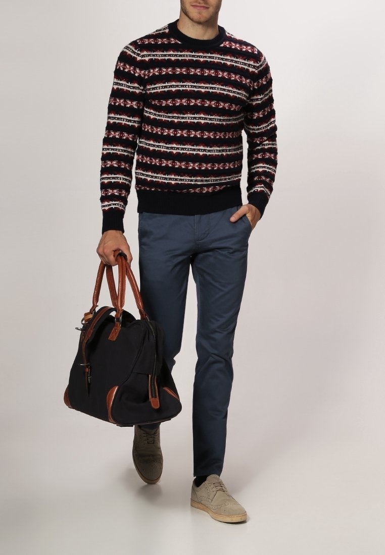 Homme portant un pull à motifs noir, blanc et rouge avec un pantalon bleu, des chaussures beiges et portant un sac noir avec des poignées marron.