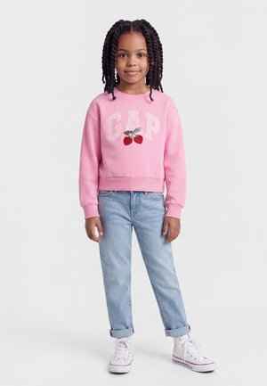 Ragazza giovane con capelli intrecciati che indossa una felpa rosa GAP con ciliegie, jeans azzurri chiari e sneakers bianche, in piedi con il volto rivolto verso lo spettatore.