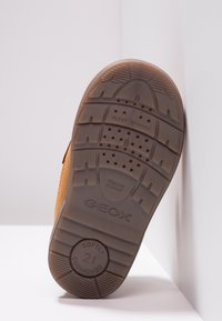 Braune Gummisohle mit strukturierten Mustern, ausgestattet mit Entwässerungsöffnungen und Branding. Der obere Bereich zeigt ein tanfarbenes Material für Langlebigkeit.