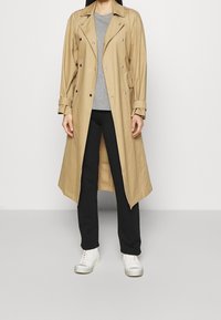 Beige Trenchcoat mit doppelter Knopfleiste, silbernen Beschlägen und Schulterklappen, getragen über einem grauen Oberteil und schwarzen Hosen, kombiniert mit weißen Sneakern.