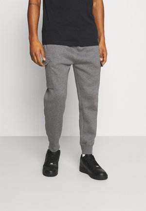 Person trägt graue Cargo-Jogginghosen, schwarze Sneakers und ein schwarzes T-Shirt und steht vor einem schlichten hellen Hintergrund.