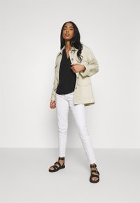 Veste oversize beige clair, t-shirt noir à col en V, jean blanc slim, et sandales noires à lanières, avec des boutons et des poches visibles.