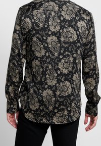 Černá košile s dlouhými rukávy s paisley a květinovým vzorem ve zlaté barvě, s rozhaleným límečkem a knoflíkovými manžetami, hladká textilie.