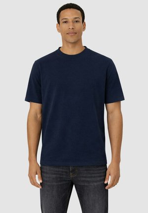 Uomo che indossa una maglietta a maniche corte blu navy semplice e jeans scuri, in piedi contro uno sfondo grigio chiaro, rivolto verso l'obiettivo.