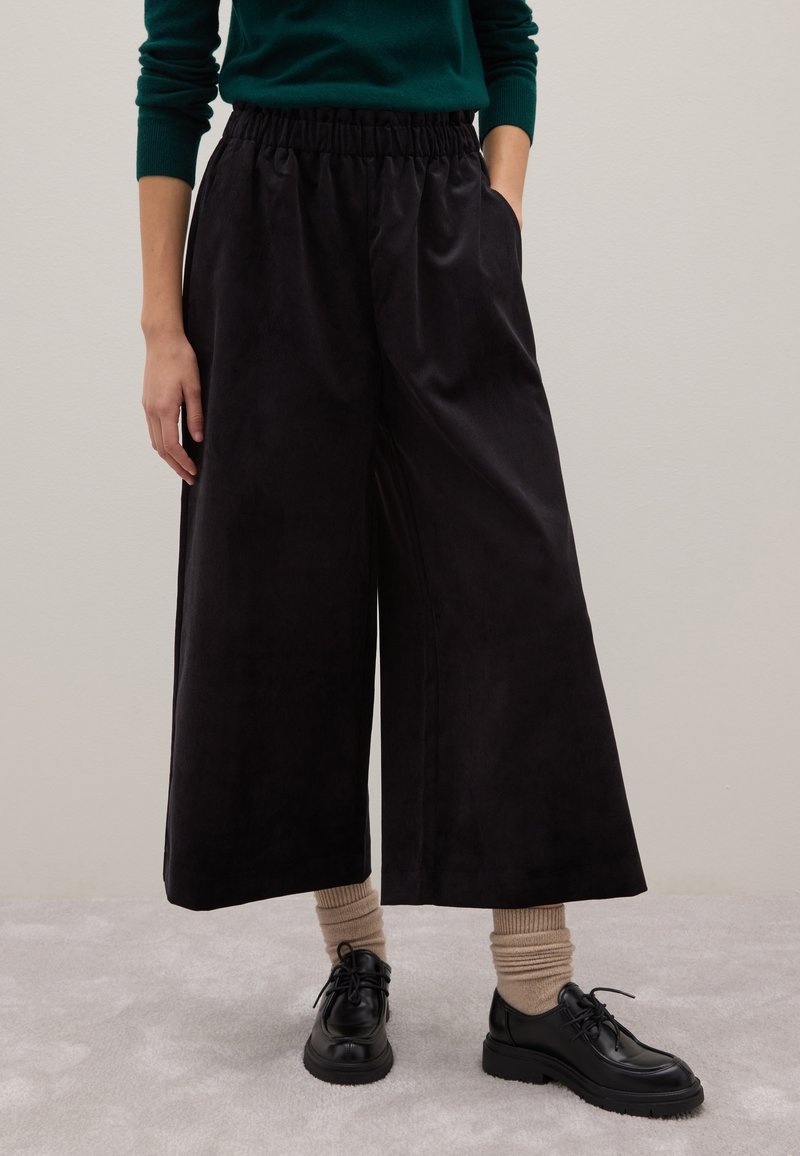 Pantaloni neri a gamba larga con una cintura elastica, realizzati in un tessuto morbido e liscio. Abbinati a scarpe nere e calzini rigati beige.