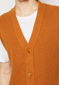 Rostfarbener gestrickter Pullover mit V-Ausschnitt, ausgestattet mit zwei Knöpfen und einem gerippten Muster, über einem weißen T-Shirt getragen.