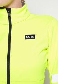 Högsynlig gul atletisk jacka med halvdragkedja, svarta detaljer och en svart GORE-logotyp på bröstet. Slät, elastisk tyg.