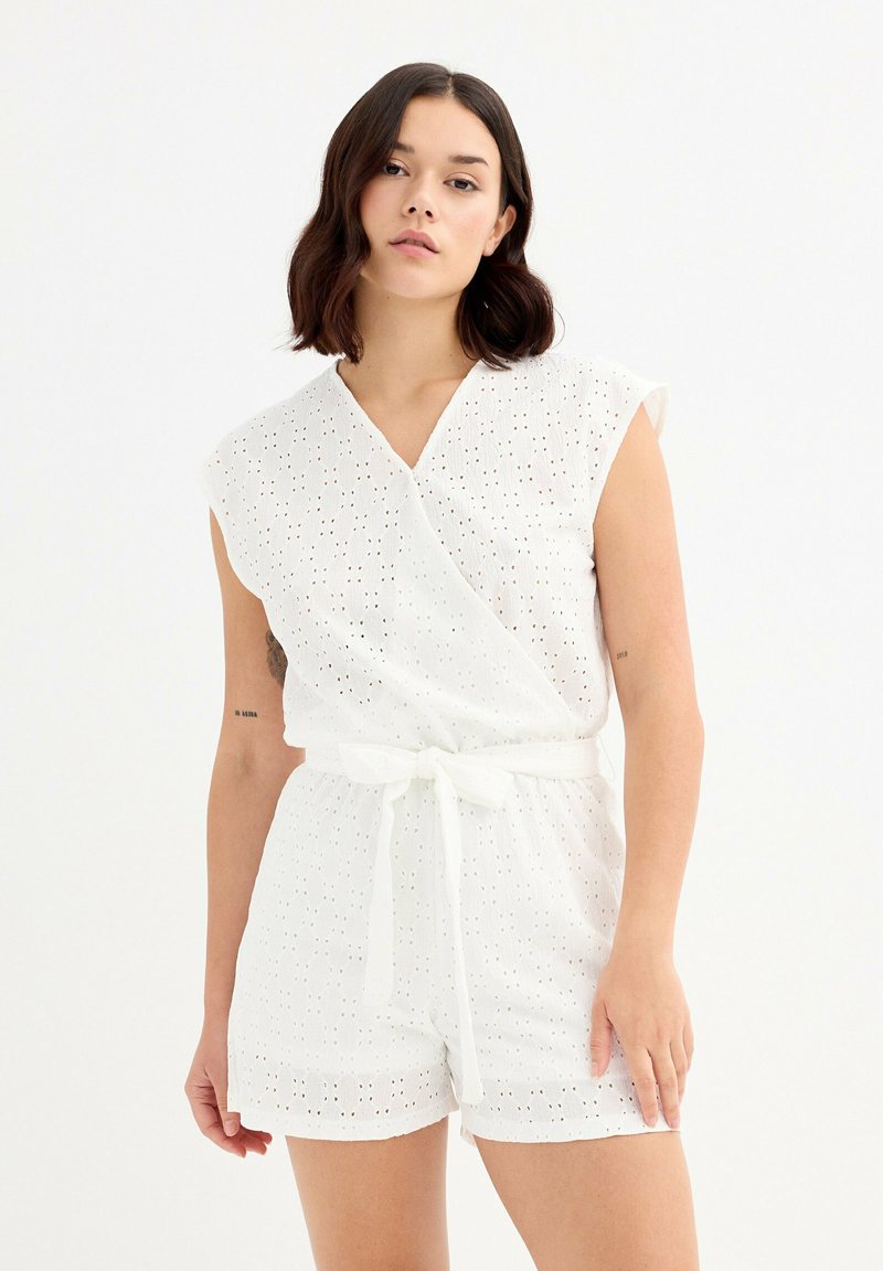 Mono de algodón blanco con diseño envolvente, mangas cortas, patrón de ojetes, cinturón atado en la cintura y pantalones cortos de corte recto. Textura suave.
