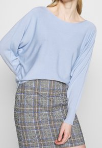 Ljusblå, löst sittande tröja med vid halsringning, kombinerad med en mönstrad rutig kjol i bruna, blå och gröna nyanser.