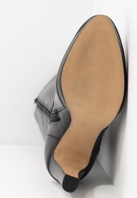 Botte en cuir noir reposant sur le côté, montrant une semelle lisse de couleur beige avec un motif discret à vagues et un petit talon bloc.