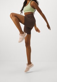 Sportovní outfit zahrnující světle zelený crop top, hnědé šortky s vysokým pasem a růžové tenisky. Výrazným detailem je logo Nike na topu.