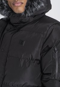 Sort puffer-jakke med en struktureret overflade, pelsforet hætte, trykknapper og en lynlåslomme på venstre arm. Logo-patch på brystet.
