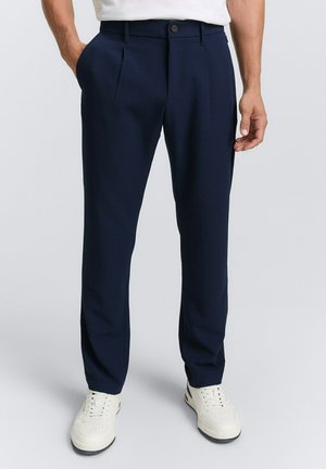 Man in marineblauwe op maat gemaakte broek en witte sneakers, met handen in de zakken, staand tegen een effen lichte achtergrond.
