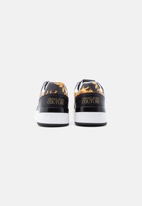 Μαύρα και χρυσά αθλητικά παπούτσια με τυπωμένο σχέδιο, λευκή σόλα, αντίθετες μαύρες λεπτομέρειες και το λογότυπο «VERSACE JEANS COUTURE» στη φτέρνα.