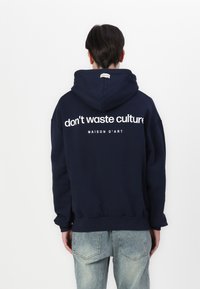 Tamno plava dukserica s kapuljačom, na kojoj se na leđima nalaze bijeli natpisi "ne troši kulturu" i "MAISON D'ART", opuštenog kroja, izrađena od mekanog materijala.