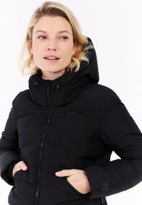 Mujer rubia vestida con una chaqueta acolchada negra con capucha, con las manos en los bolsillos, mirando ligeramente hacia su izquierda contra un fondo blanco.