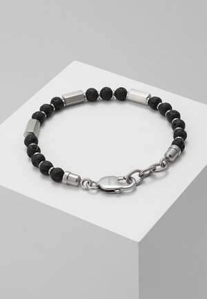 Bracelet avec perles en pierre de lave noire et accents cylindriques en argent, comprenant un fermoir homard argenté sur un piédestal blanc.