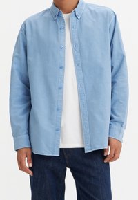 Chemise bleu clair en coton avec col pointu, manches longues et coupe décontractée, portée sur un T-shirt blanc.