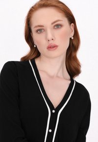 Pulover negru cu margini albe, de tip V, cu nasturi asemănători perlului. Are mâneci lungi și o textură moale. Design simplu, clasic.