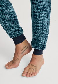 Geprinte joggers in donkerblauw en teal hebben een geometrisch driehoekpatroon, met elastische boorden bij de enkels. Blote voeten zijn zichtbaar, met tattoos.
