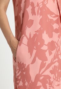 Ärmelloses Kleid aus weichem pinken Stoff, mit einem Blumenmuster in einem dunkleren Pinkton. Verfügt über eine Seiten pocket für praktische Verwendung.