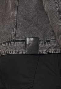 Zwarte denim met een gestructureerde afwerking, voorzien van een leren label met geperste tekst nabij de zoom. Eenvoudig ontwerp, minimale hardware.