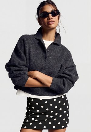 Femeie purtând ochelari de soare negri, pulover gri închis cu nasturi, cămașă albă dedesubt și fustă neagră cu buline albe, cu brațele încrucișate.