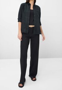 Chemise en lin noire avec manches retroussées, boutonnage sur le devant et une poche poitrine unique, associée à un pantalon noir à jambes larges et des sandales noires à enfiler.