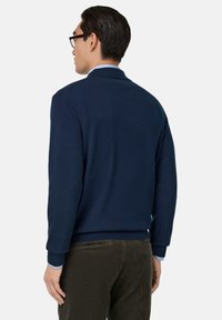 Navyblauer Strickpullover mit einem gerippten Kragen und Bündchen, der eine subtile Textur aufweist. Über einem hellblauen Hemd getragen und mit dunklen Hosen kombiniert.