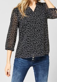Blouse noire transparente avec des pois gris, décolleté en V et manches trois-quarts, assortie à un jean en denim bleu.
