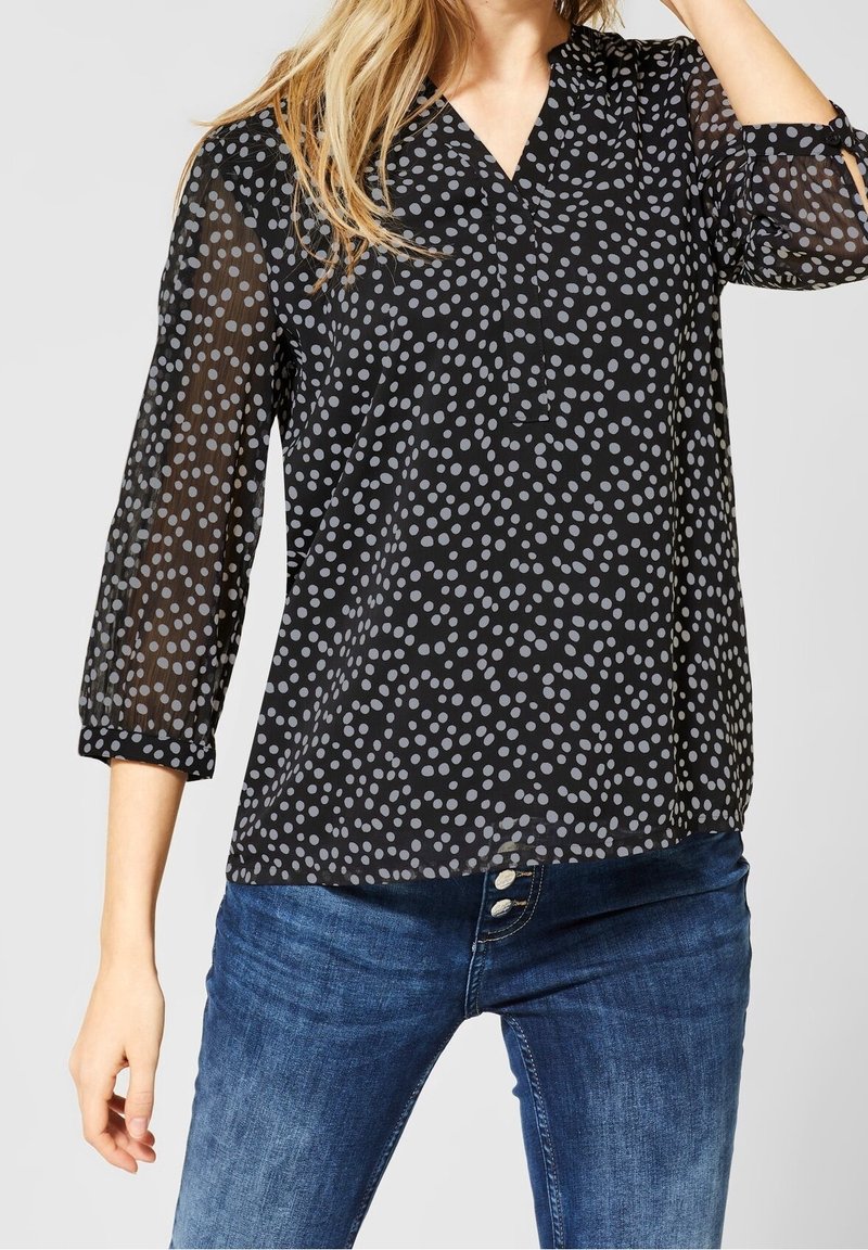 Blouse noire transparente avec des pois gris, décolleté en V et manches trois-quarts, assortie à un jean en denim bleu.