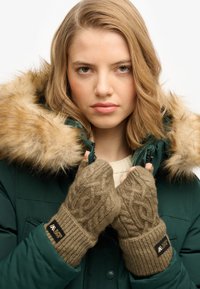 Jonge vrouw in een groene winterjas met een bontkraag, die de jascol met beide handen vasthoudt en bruin gebreide vingerloze handschoenen draagt.
