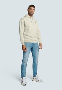 Lichtbeige hoodie met een kangoeroezak en een trekkoord, gecombineerd met lichtblauwe jeans en witte sneakers met zwarte accenten.