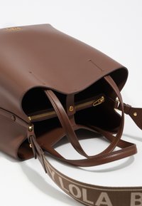Bolso de cuero marrón con un diseño estructurado, que cuenta con un bolsillo con cremallera dorada en el interior y un logotipo en la parte frontal. Correa desmontable con detalle de texto.