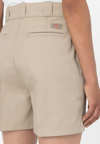 Béžové šortky vyrobené z pevného materiálu, s stredne vysokým pásom, dvoma zadnými vreckami a malou nášivkou s logom na zadnej strane.