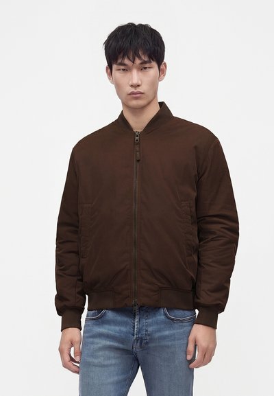 Jeune homme portant une veste bomber marron foncé zippée et un jean bleu clair sur un fond blanc uni.