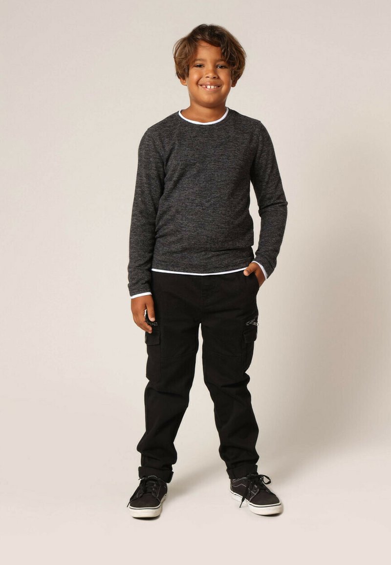 Garçon souriant avec des cheveux bruns debout, portant un t-shirt à manches longues gris foncé, un pantalon cargo noir et des baskets noires avec des semelles blanches.