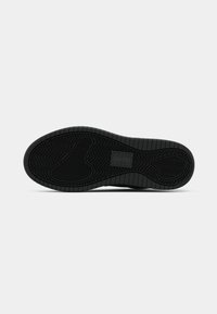 Semelle d'une basket noire avec des motifs de bande de roulement texturés et une zone rectangulaire pour le logo au centre sur un fond clair uni.