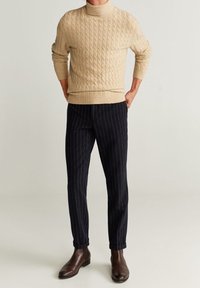 Homme portant un pull à col roulé beige en maille torsadée, un pantalon bleu marine à rayures fines, et des bottines en cuir marron foncé, debout les mains dans les poches.