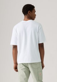 White cotton t-shirt with a relaxed fit and short sleeves, viewed from the back. Accompanied by light green cargo pants and textured details.