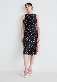 Zwarte jurk met een verspreid bloempatroon, grote strikdetail bij de halslijn, aangesloten silhouet, gecombineerd met een roze handtas en platform schoenen.