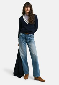 Maglione blu navy a scollo a V con bordo bianco, abbinato a jeans dritti azzurri chiari e stivaletti alla caviglia in camoscio marrone. Una giacca blu navy appoggiata su un braccio.