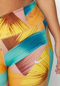 Leggings de cintura alta em padrão vibrante de folhas de palmeira multicoloridas; apresenta tons de azul-piscina, amarelo e castanho com logo discreto da Nike e tecido elástico.
