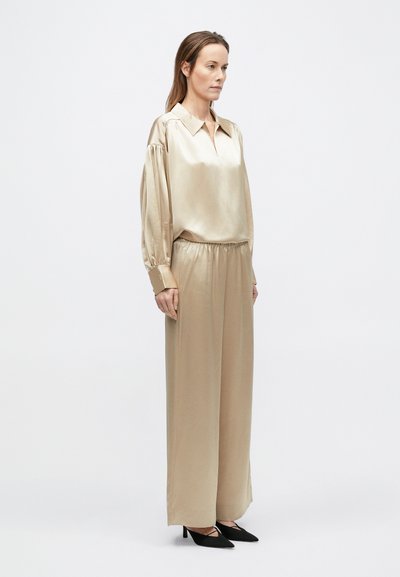 Nő áll, laza bézs szatén blúzban, hosszú puffos ujjal és hozzáillő, bő szárú nadrágban, fekete hegyes sarkú cipővel párosítva.