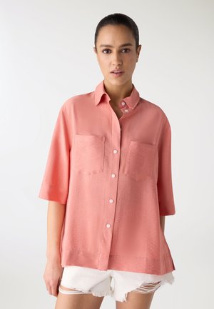 Donna con capelli raccolti che indossa una camicia corallo ampia con due tasche sul petto e pantaloncini bianchi strappati, su sfondo bianco.