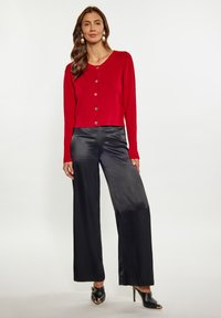 Rode, knoopsluiting, cropped cardigan met lange mouwen, gecombineerd met zwarte, wijde satijnen broek en zwarte open-teenschoenen. Effen achtergrond.